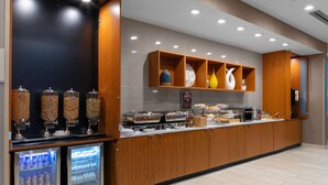 Una barra de desayuno moderna con dispensadores refrigerados de bebidas, diversas opciones de comida y estantes decorativos.