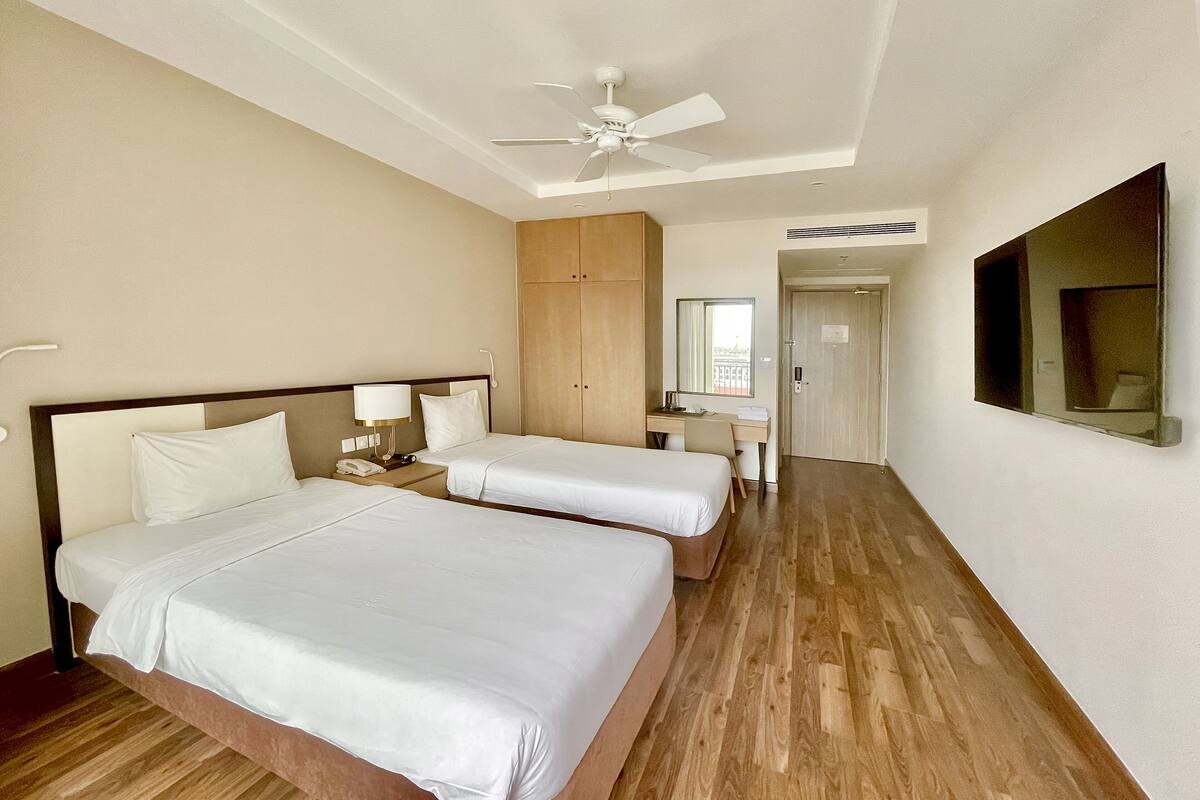 A hotel room with two beds, a desk, a wardrobe, and a ceiling fan.
