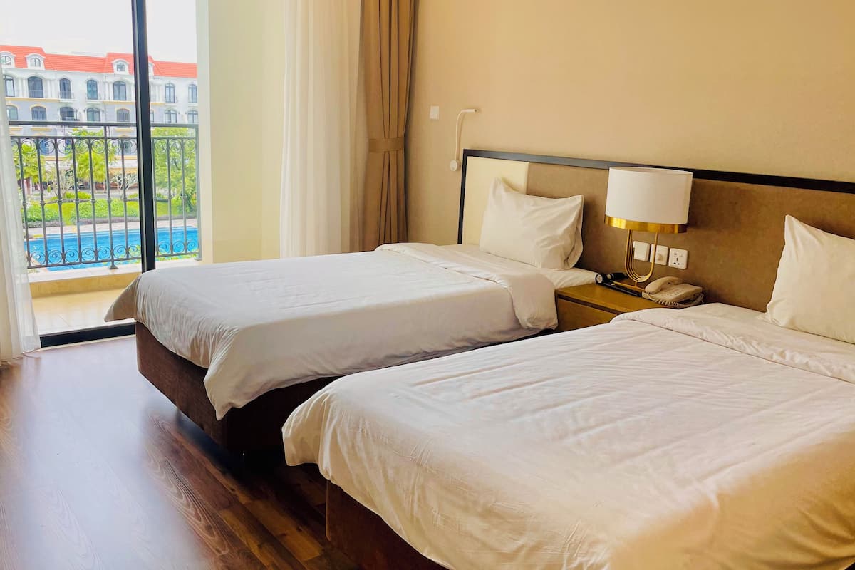 A hotel room with two beds, a nightstand with a phone, and a view of a building and pool through the window.