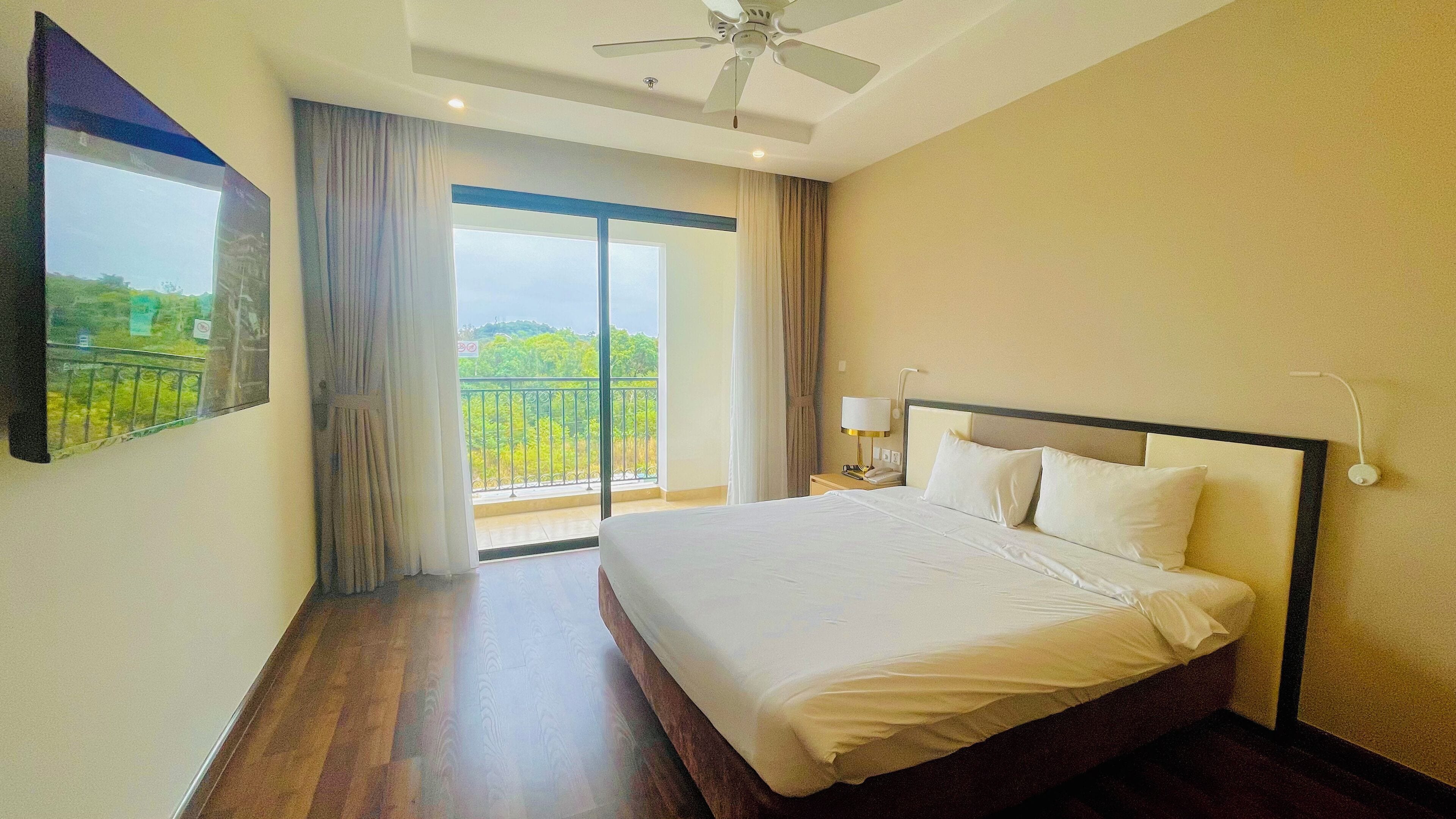 Quarto de hotel com uma cama grande, um ventilador de teto, mesa de cabeceira com abajur, vista para o verde pela janela e uma varanda.