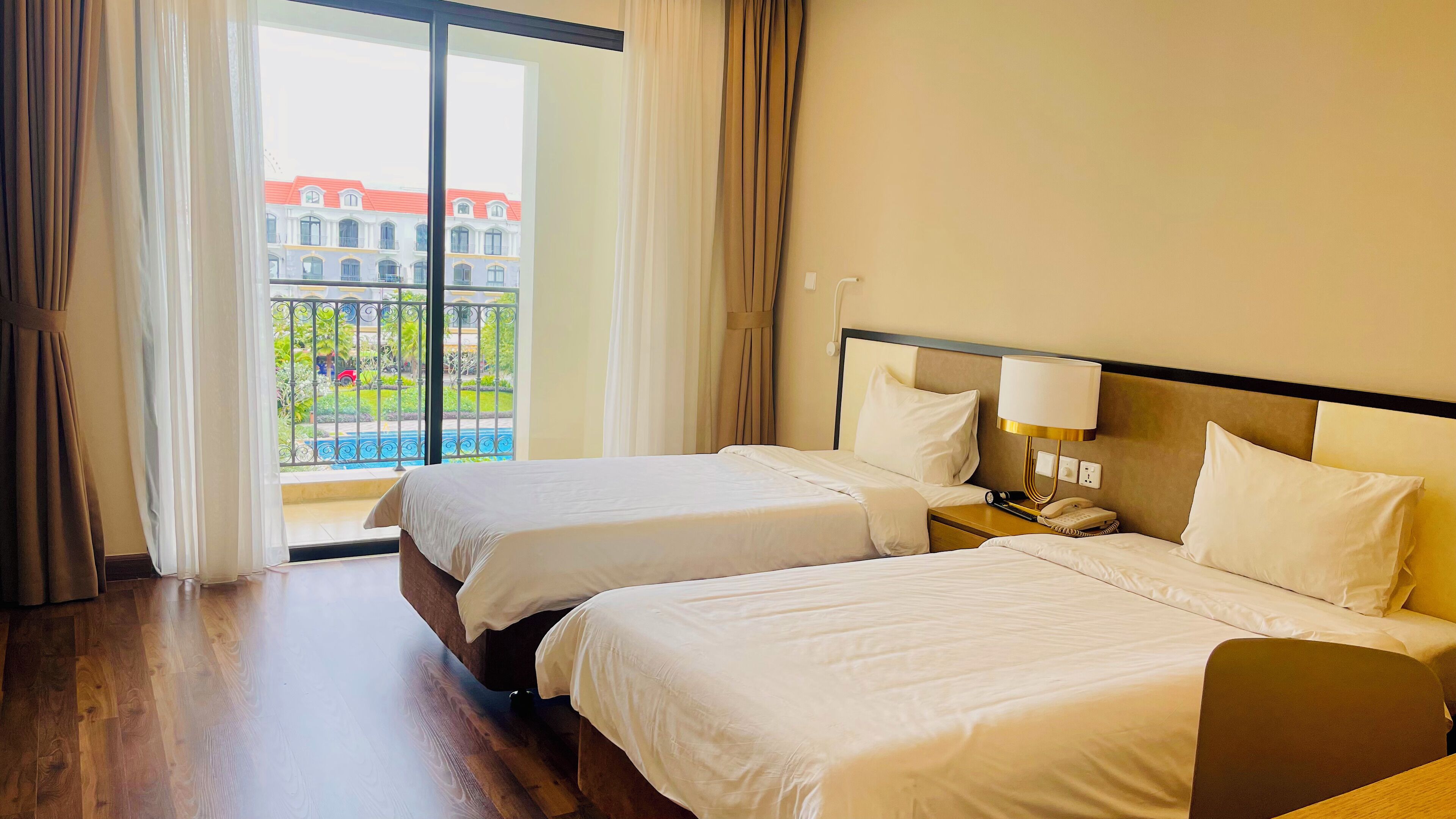 Habitación de hotel con dos camas, una mesita de noche con lámpara y vistas a un edificio y zonas verdes a través de la ventana.