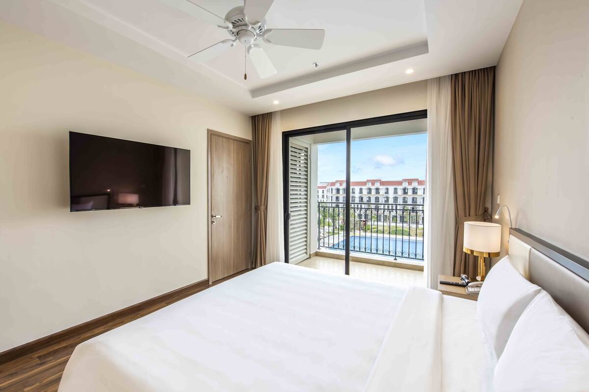 A hotel room with a large bed, a ceiling fan, a TV mounted on the wall, and a balcony with a view of buildings.
