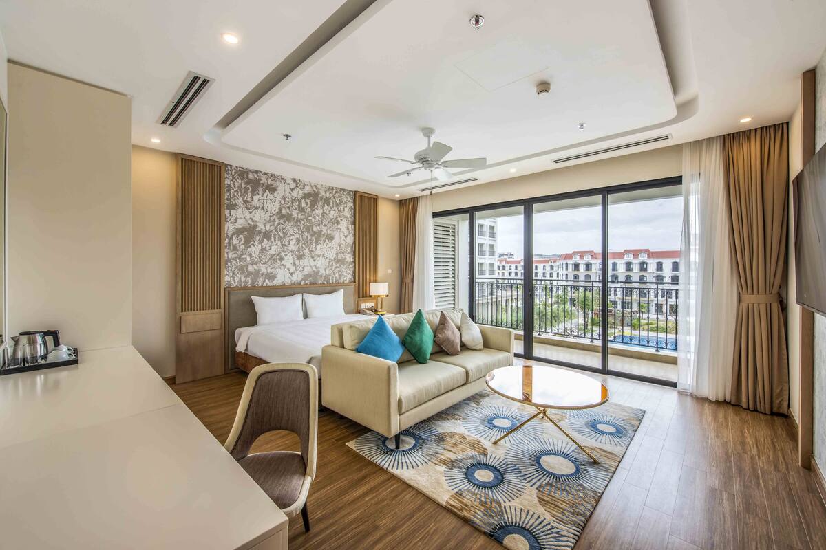 A hotel room with a bed, a sofa, a desk, a chair, a TV, and a large window with a view of buildings.