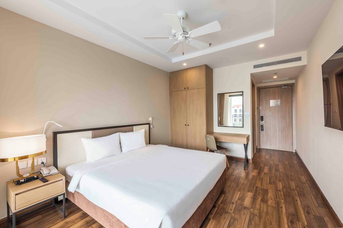 A hotel room with a bed, bedside table, desk, chair, wardrobe, and a ceiling fan.
