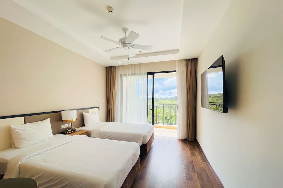 A hotel room with two beds, a ceiling fan, a TV, and a balcony with a view of greenery.