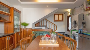 A modern kitchen with wooden cabinets, a dining table with chairs, a staircase, and a washing machine. - Chipa Homestay (Da Nang)