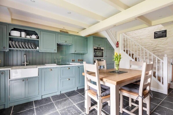 A kitchen with teal cabinets, a wooden dining table, and a staircase with a white railing. - 2 Bedroom Cottage Close to Perranporth Beach (Perranzabuloe)
