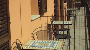 Un étroit couloir extérieur avec des chaises et des tables en métal, un sol carrelé et un bâtiment doté de volets.