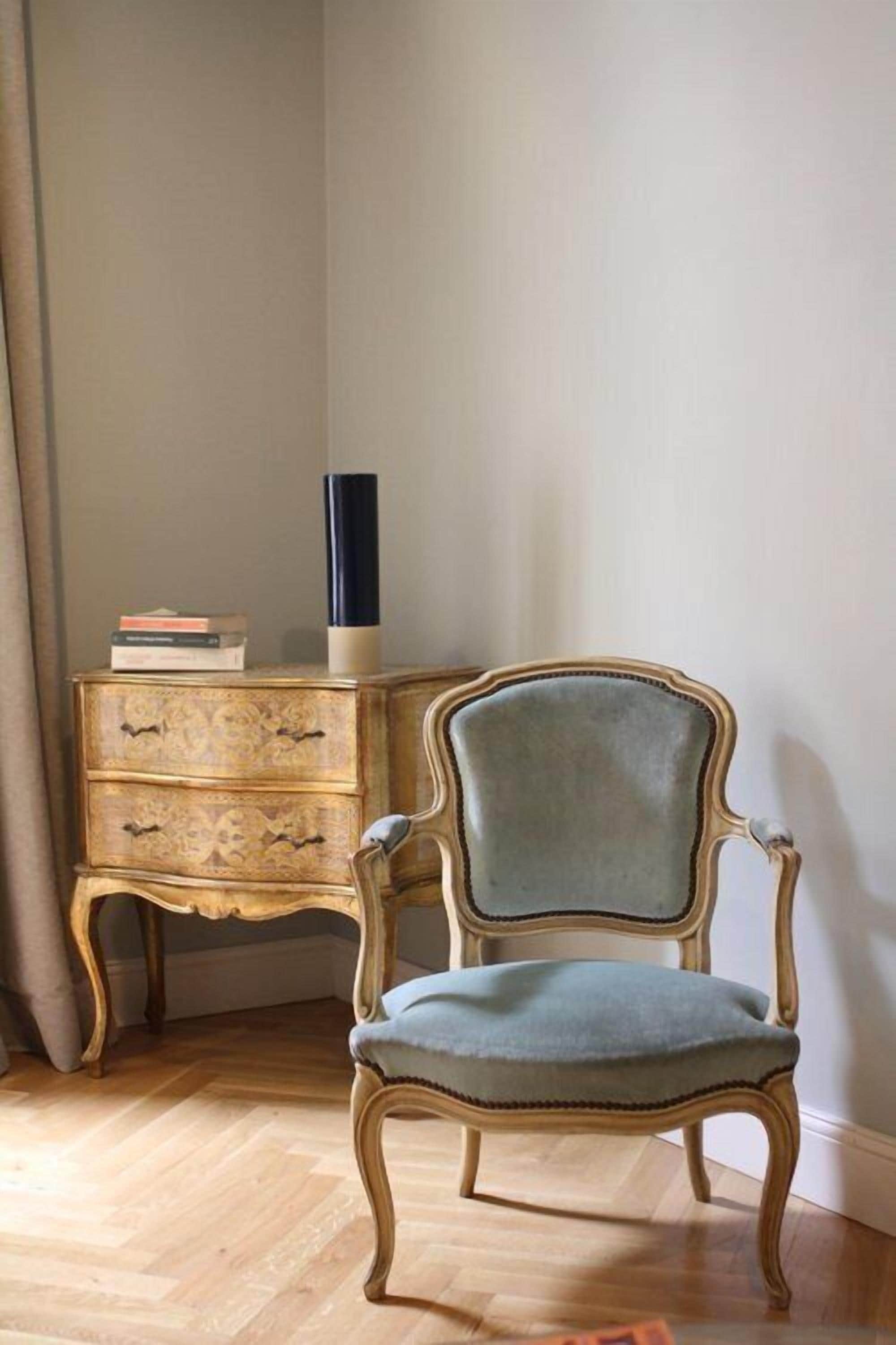 Een kamer met een stoel in vintage stijl, een houten dressoir met lades, en een hoge vaas erop.