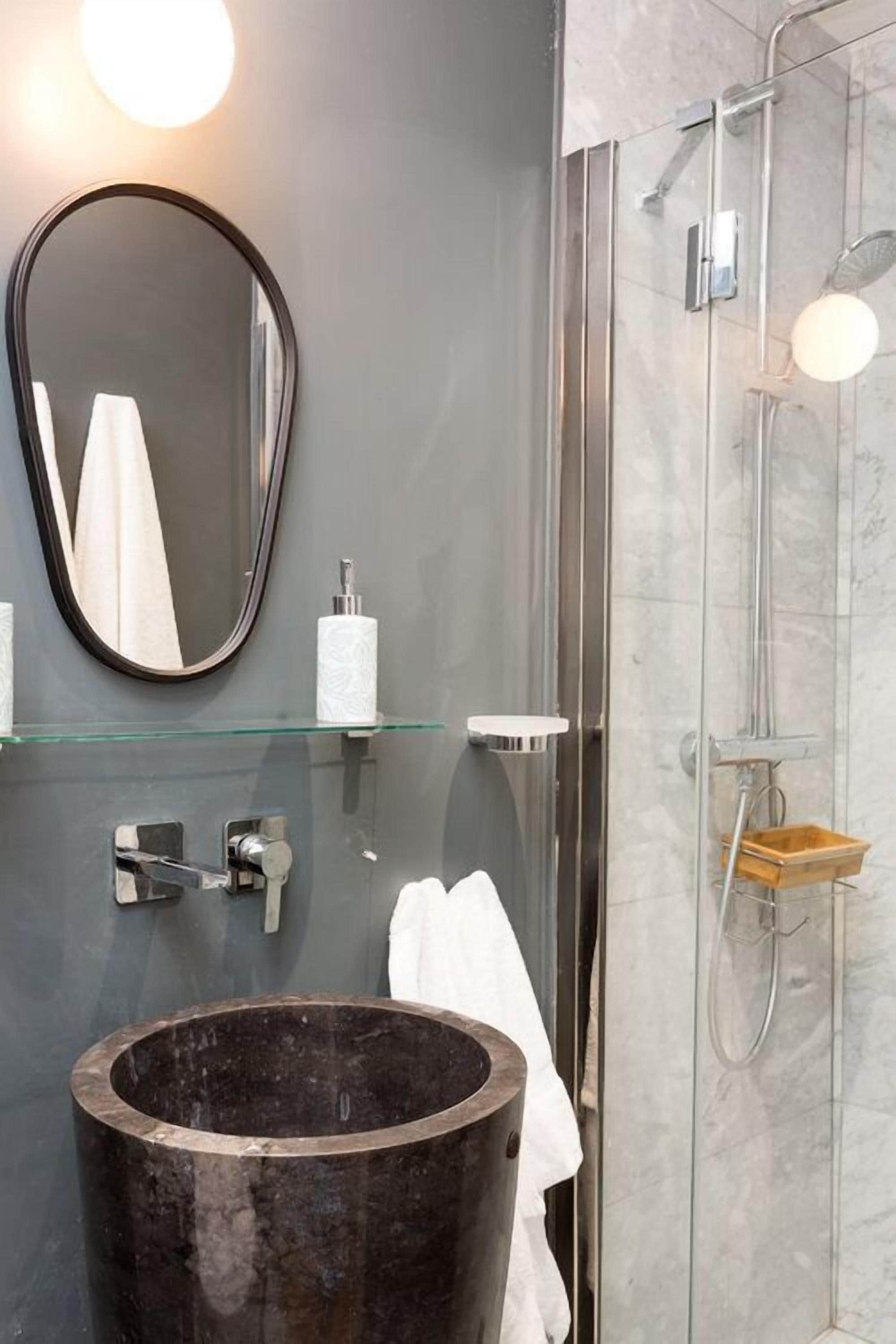 Un baño moderno con un espejo ovalado grande, un lavabo redondo, un estante de vidrio con artículos de aseo y una zona de ducha.