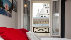 A bedroom with a bed, a red pillow, a white pillow, and a view of a white building through a window with a black metal gate.
