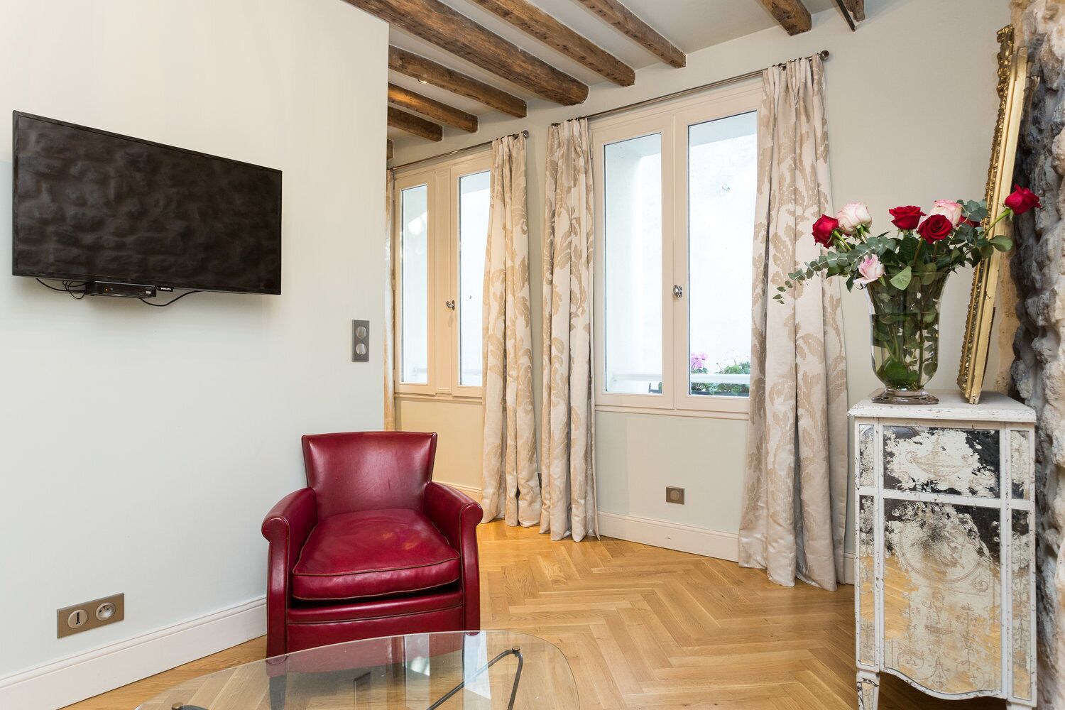 Una habitación con piso de madera, un sillón de cuero rojo, una mesa de vidrio, un televisor montado en la pared y una ventana con cortinas.