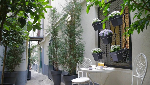 A narrow outdoor corridor with potted plants, a round table with chairs, and hanging flower baskets.