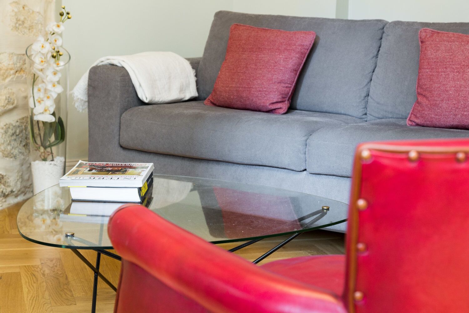 Un salon avec un canapé gris, des coussins rouges, une table basse en verre sur laquelle repose un livre, et un fauteuil rouge.