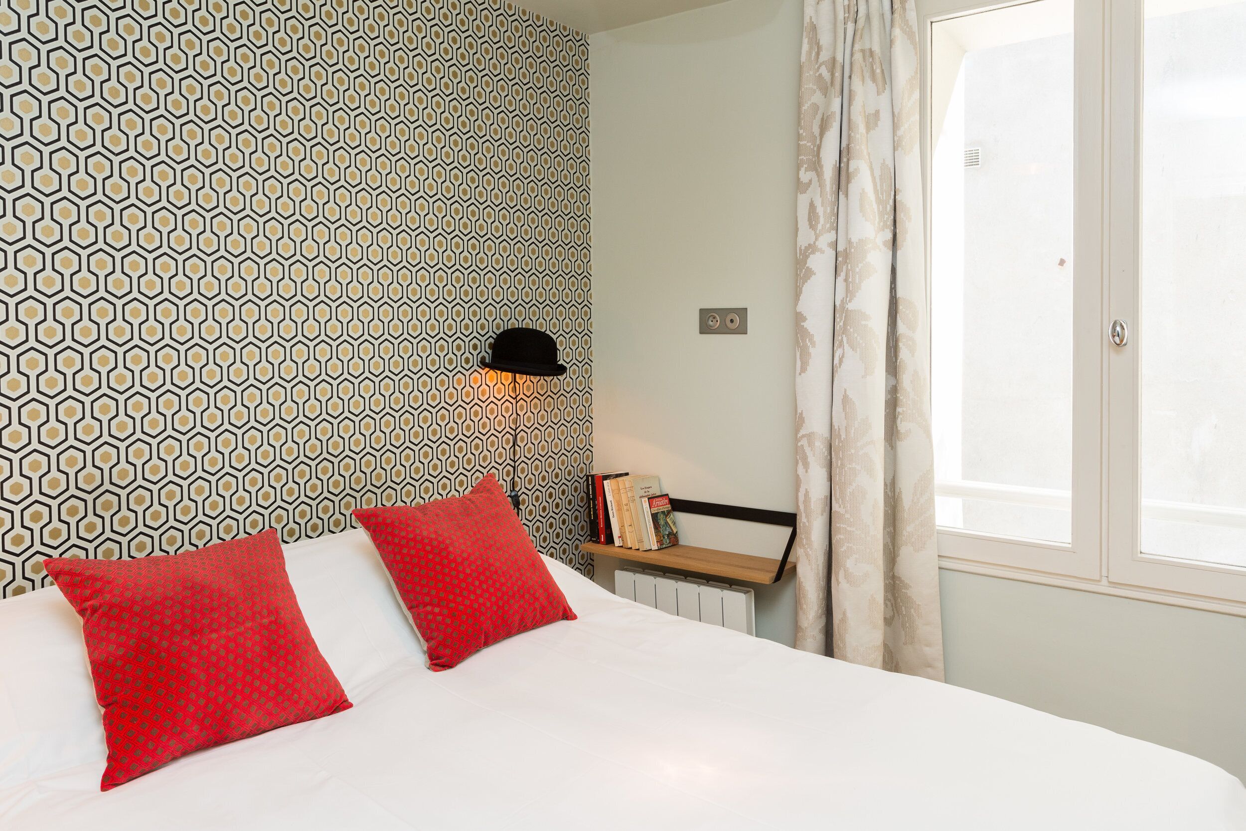 A neatly made bed with red pillows, a patterned wallpaper, a black hat on a shelf, and a window with curtains.