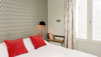 A neatly made bed with red pillows, a patterned wallpaper, a black hat on a shelf, and a window with curtains.