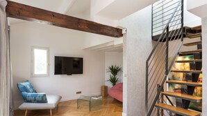 A modern living room with a wooden ceiling beam, a TV mounted on the wall, a glass coffee table, a blue patterned sofa, a red sofa, and a staircase with a metal railing.