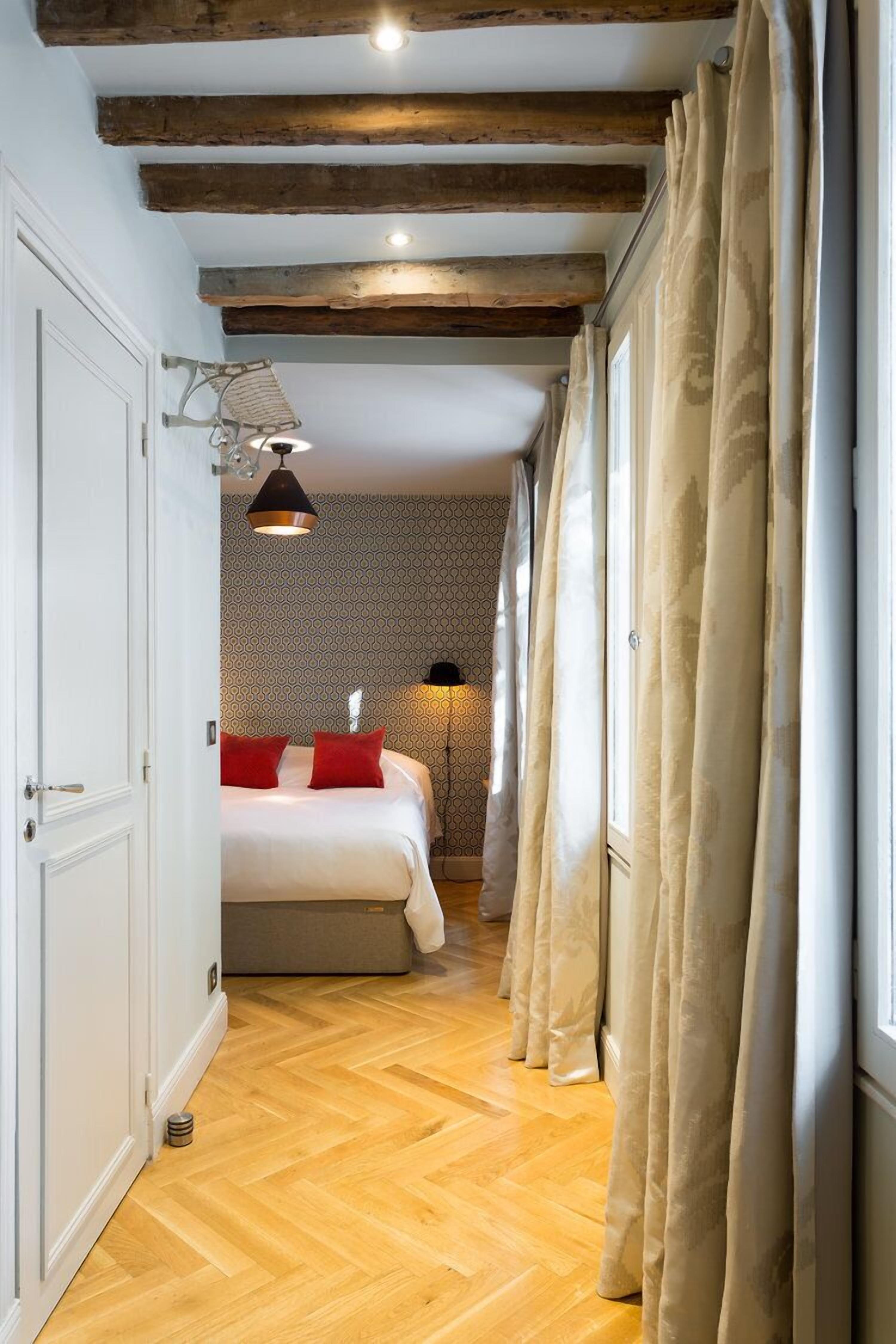 A narrow hallway leading to a bedroom with a bed, a hanging light fixture, and a window with curtains.