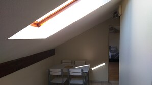 Un petit coin repas avec une table et quatre chaises, une lucarne au-dessus et un lit à l’arrière-plan.