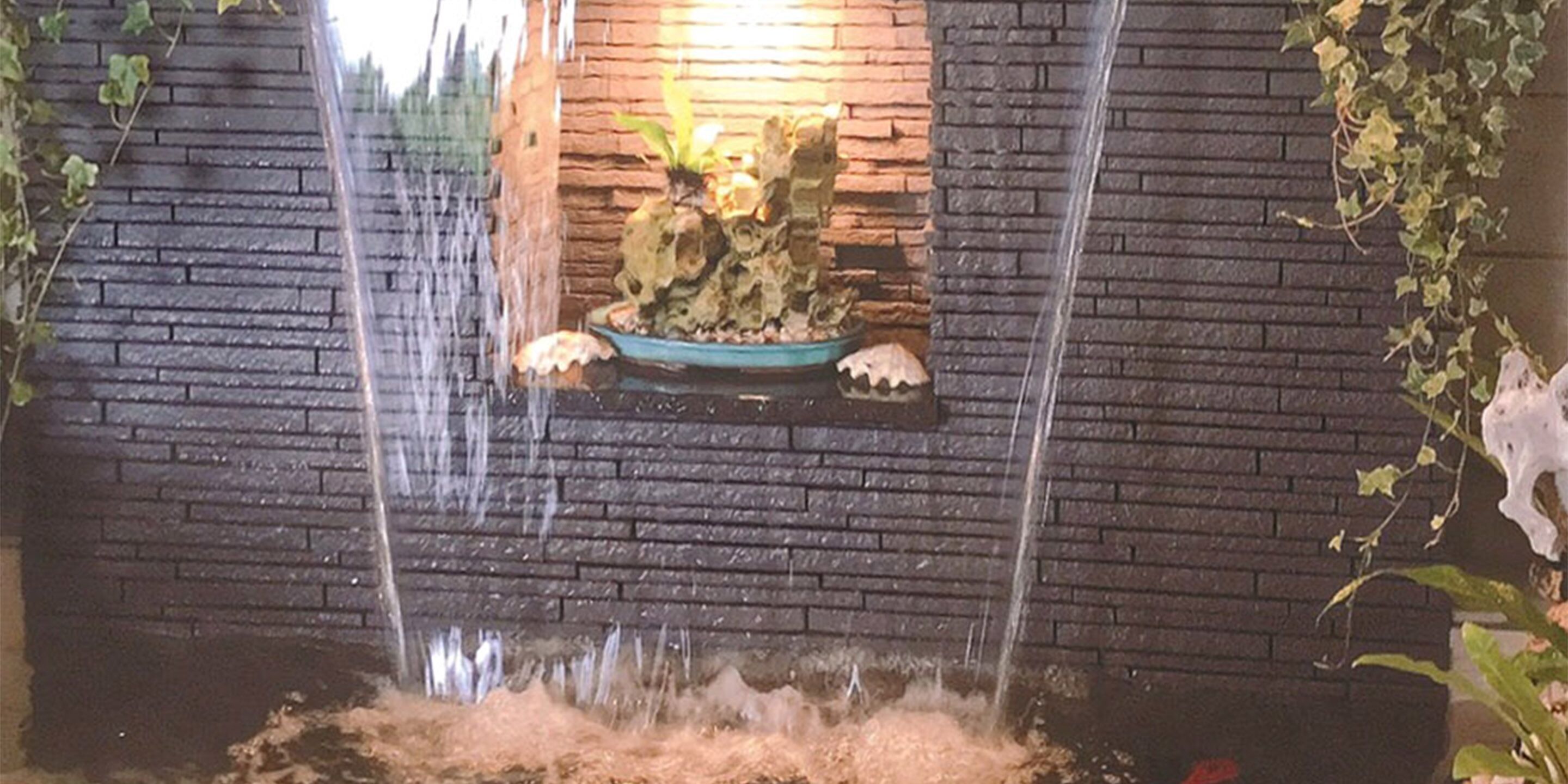Ein Wasserfall mit einem steinernen Becken in der Mitte, umgeben von einer Ziegelmauer und Grünpflanzen.