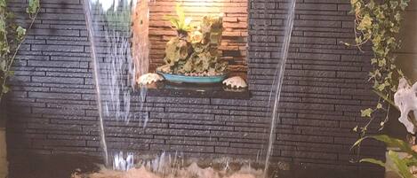 A waterfall feature with a stone basin in the center, surrounded by a brick wall and greenery.