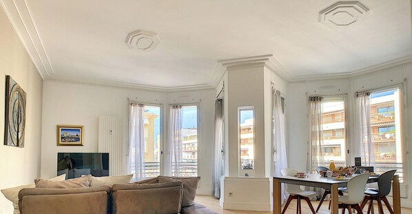 Uma sala de estar com sofá, área de jantar com mesa e cadeiras, e grandes janelas com varandas visíveis do lado de fora.