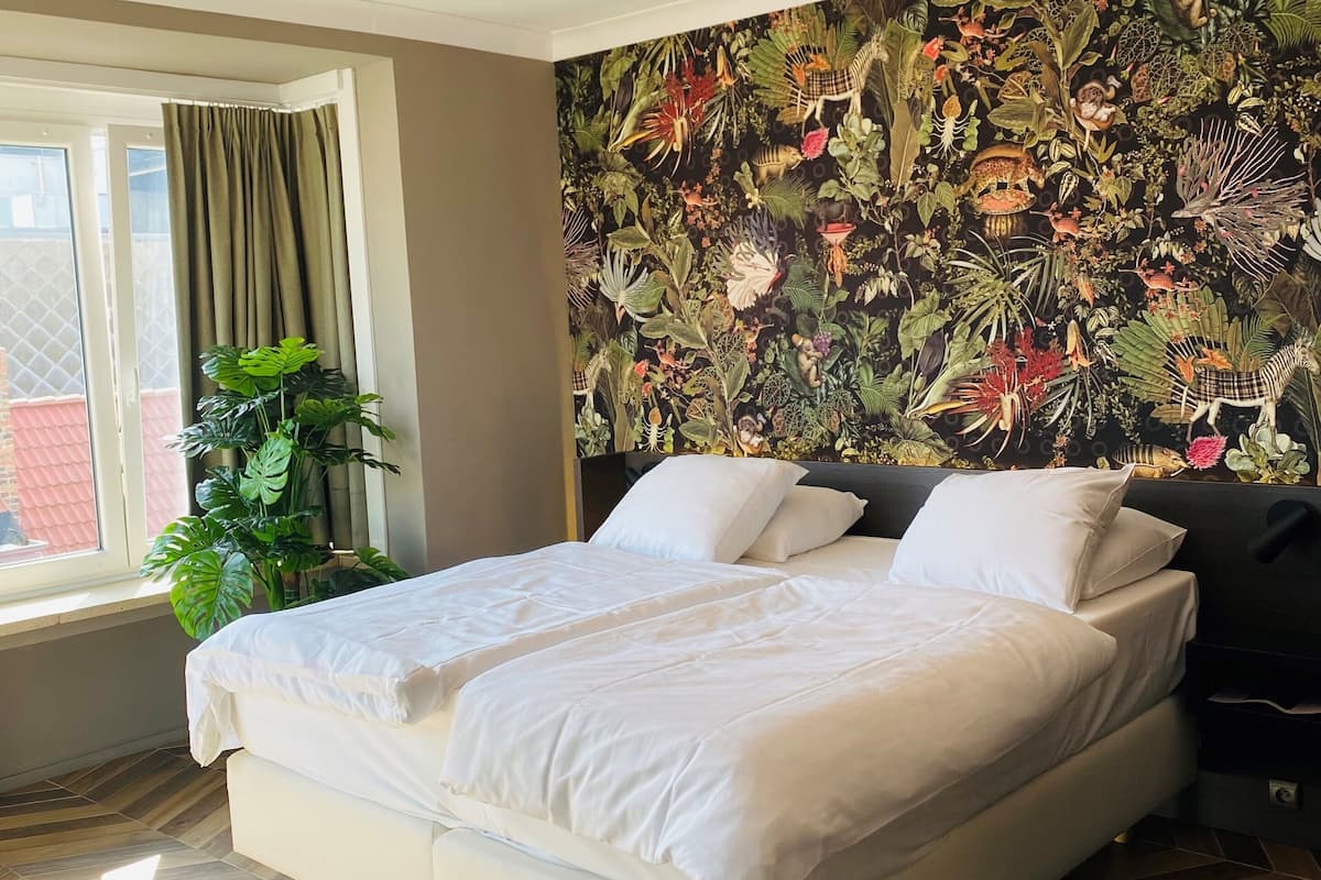Un dormitorio con una cama, un ventanal grande, una planta y un papel tapiz con motivos selváticos.