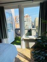 A room with a large window, a view of the city, a small balcony, and a potted plant.