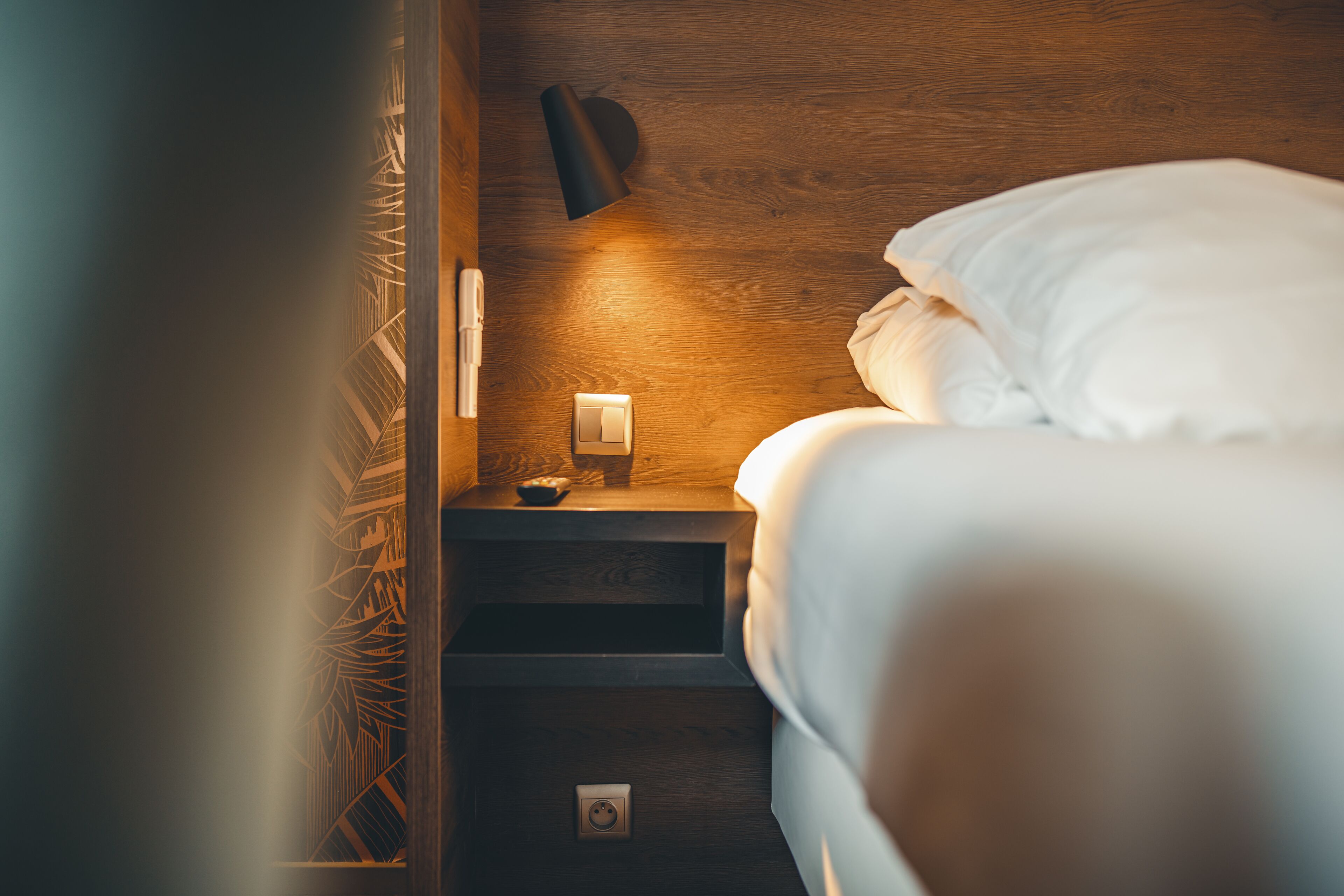 Un lit bien fait, avec du linge de lit blanc, une lampe de chevet et une prise électrique sur une table de chevet en bois.