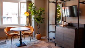 A modern living room with a round table, two orange chairs, a potted plant, a TV mounted on a wooden cabinet, and a large window with curtains. - Prado Next Door (Ostend)