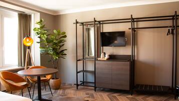 Habitación de hotel con cama, una mesa redonda con sillas, una planta en maceta, un televisor montado en la pared y una estantería metálica.