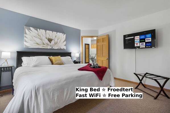 Una cama tamaño king con una manta roja, un televisor de montaje en pared que muestra servicios de streaming, y una silla independiente.