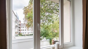 Een raam met twee witte kaarsen op een houten dienblad, uitkijkend op een straat met gebouwen en bomen.
