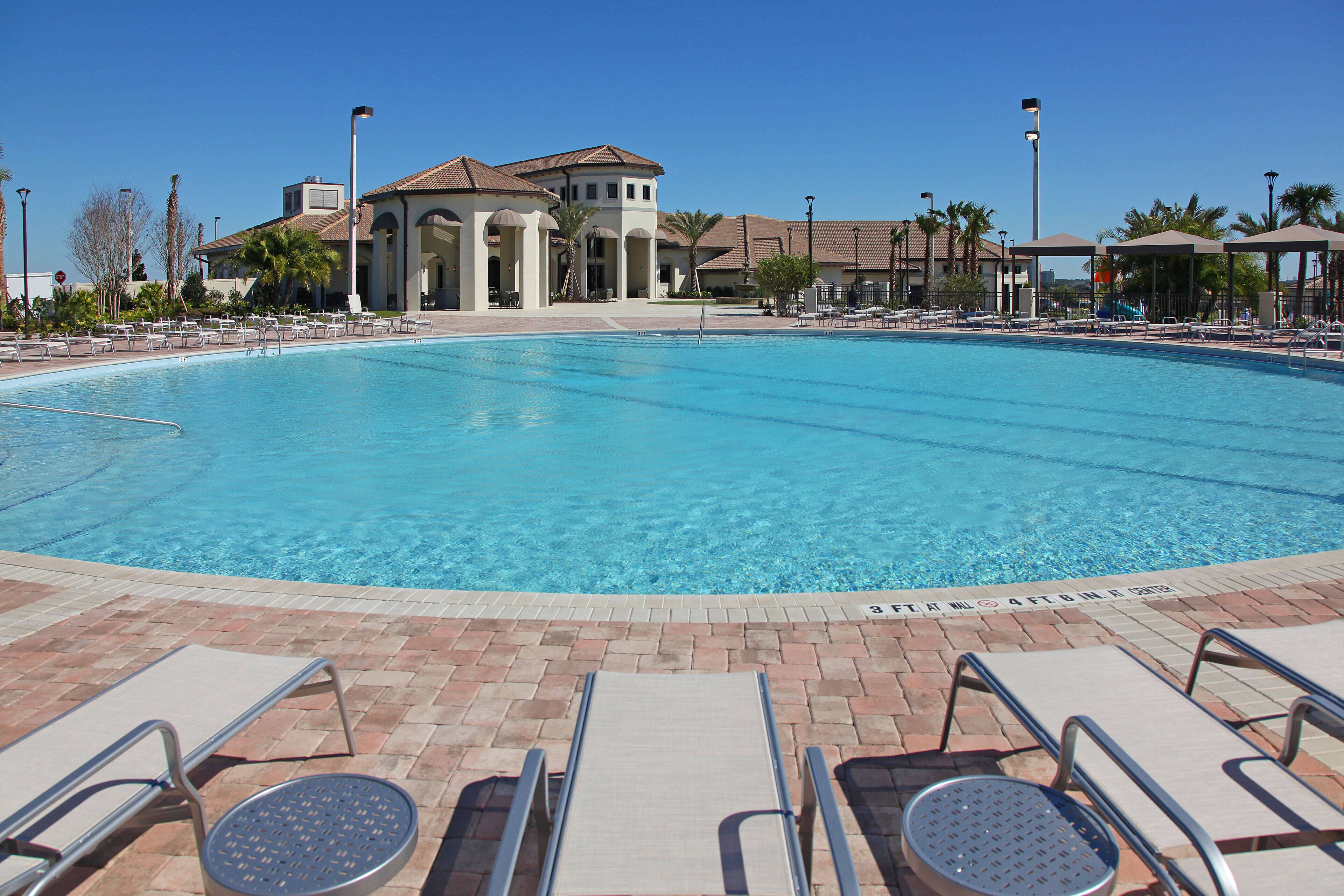 A large swimming pool with lounge chairs and umbrellas, surrounded by buildings with a tiled roof and palm trees.
