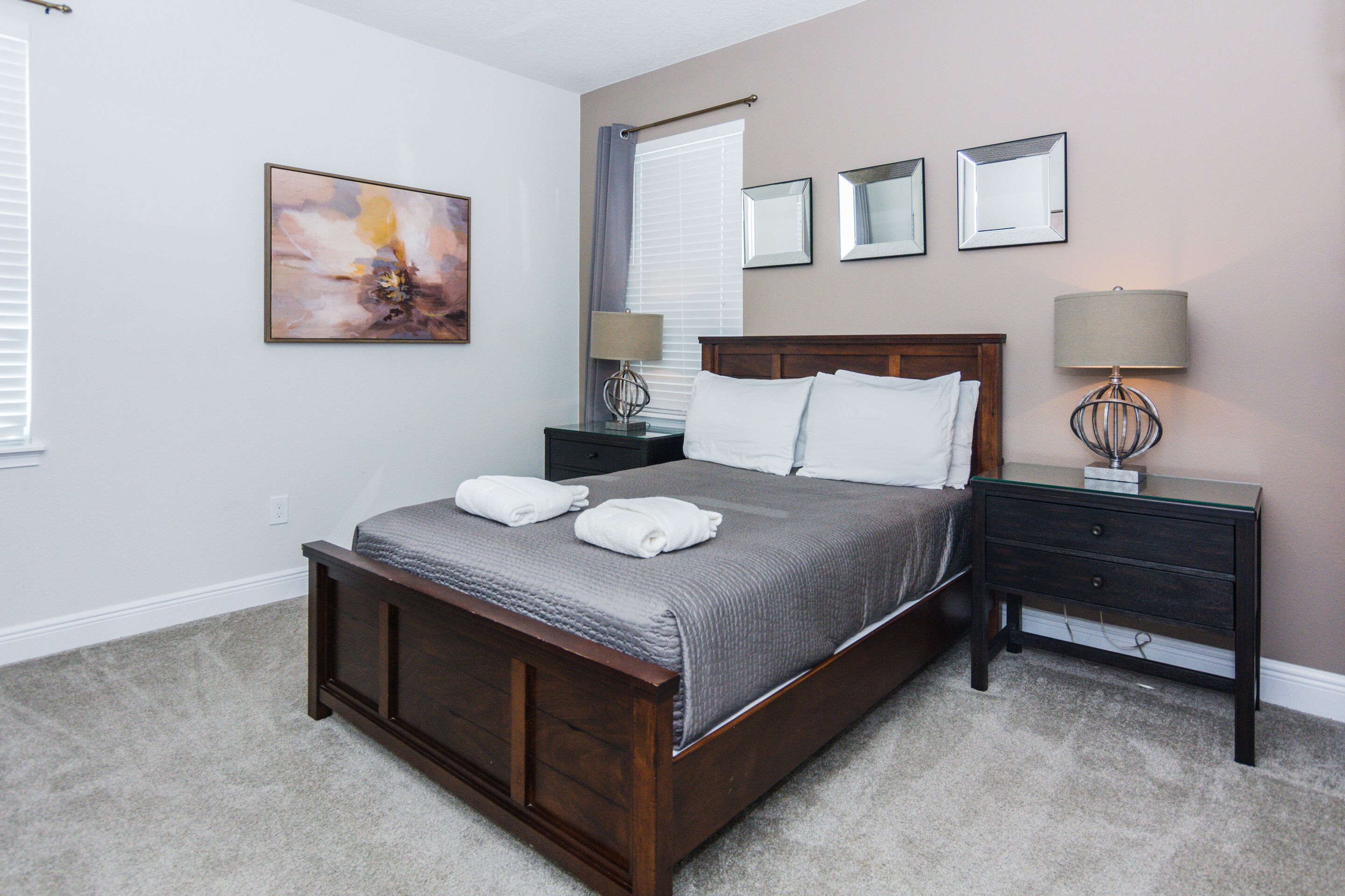 A bedroom with a wooden bed, two bedside tables with lamps, a window with curtains, and a painting on the wall.