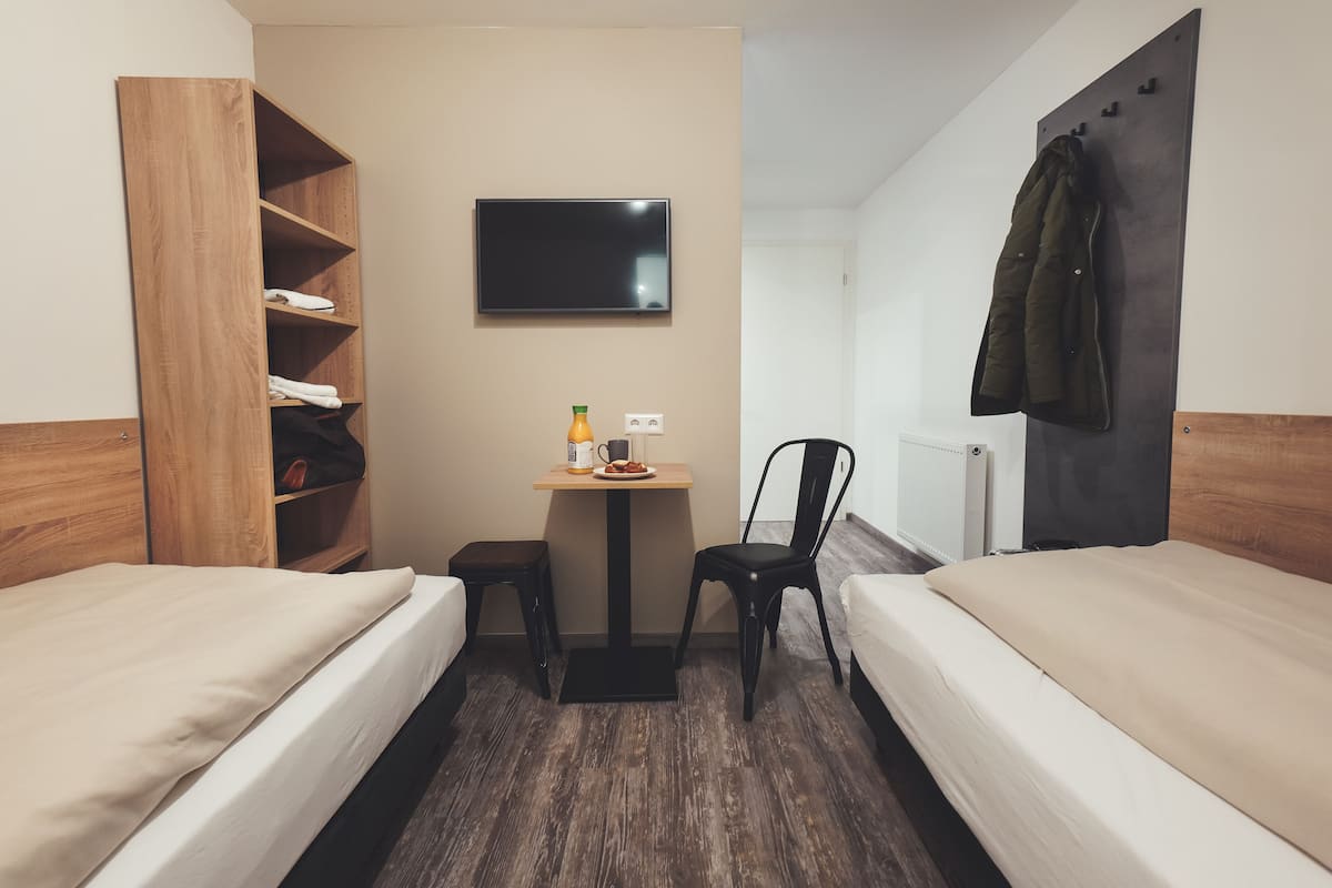 Une chambre d’hôtel avec deux lits, une armoire en bois, une petite table avec une bouteille et une assiette, un téléviseur fixé au mur et un manteau accroché à un crochet.