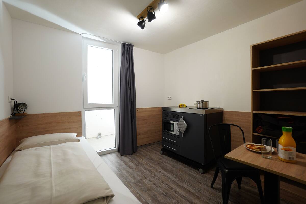 Une chambre d’hôtel comprenant un lit, un bureau avec une chaise, un four à micro-ondes et une étagère avec une bouteille.