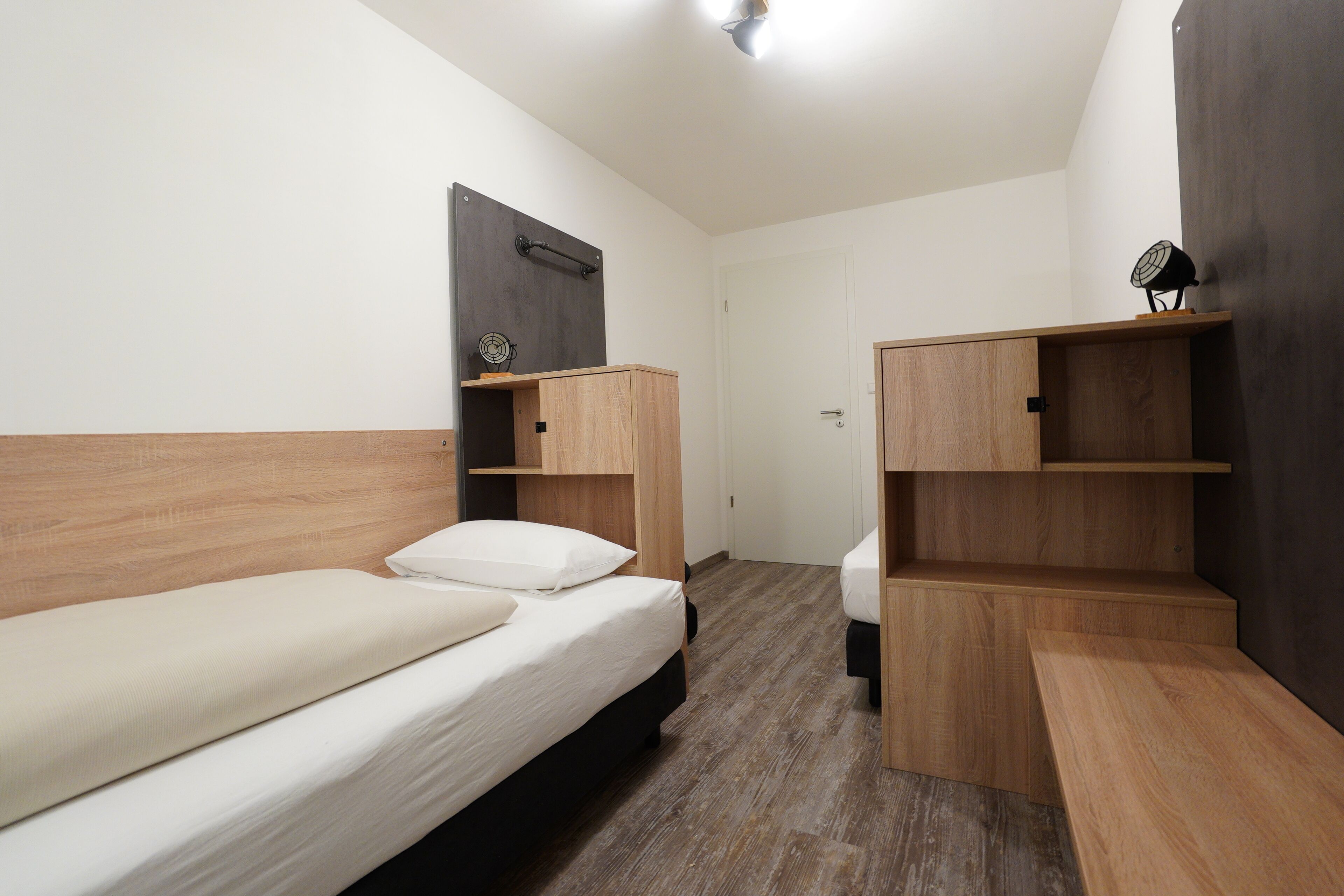 Una habitación pequeña y moderna con dos camas individuales, cabeceros de madera y estantes empotrados.