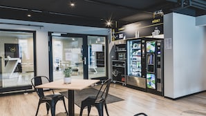 Un interior moderno con una mesa y sillas, una máquina expendedora y puertas de vidrio que dan a otra zona.