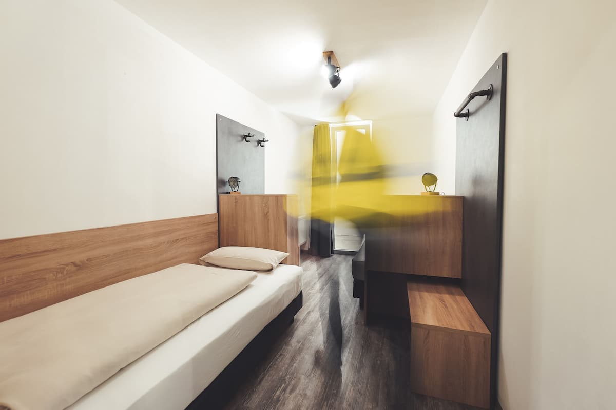 Une chambre de taille modeste, comprenant un lit simple, une tête de lit en bois, une table de chevet, un miroir et un petit meuble en bois.