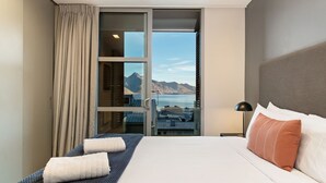 Una camera d'hotel con un letto, un comodino, una lampada e vista sulle montagne e sull'acqua attraverso una porta scorrevole.