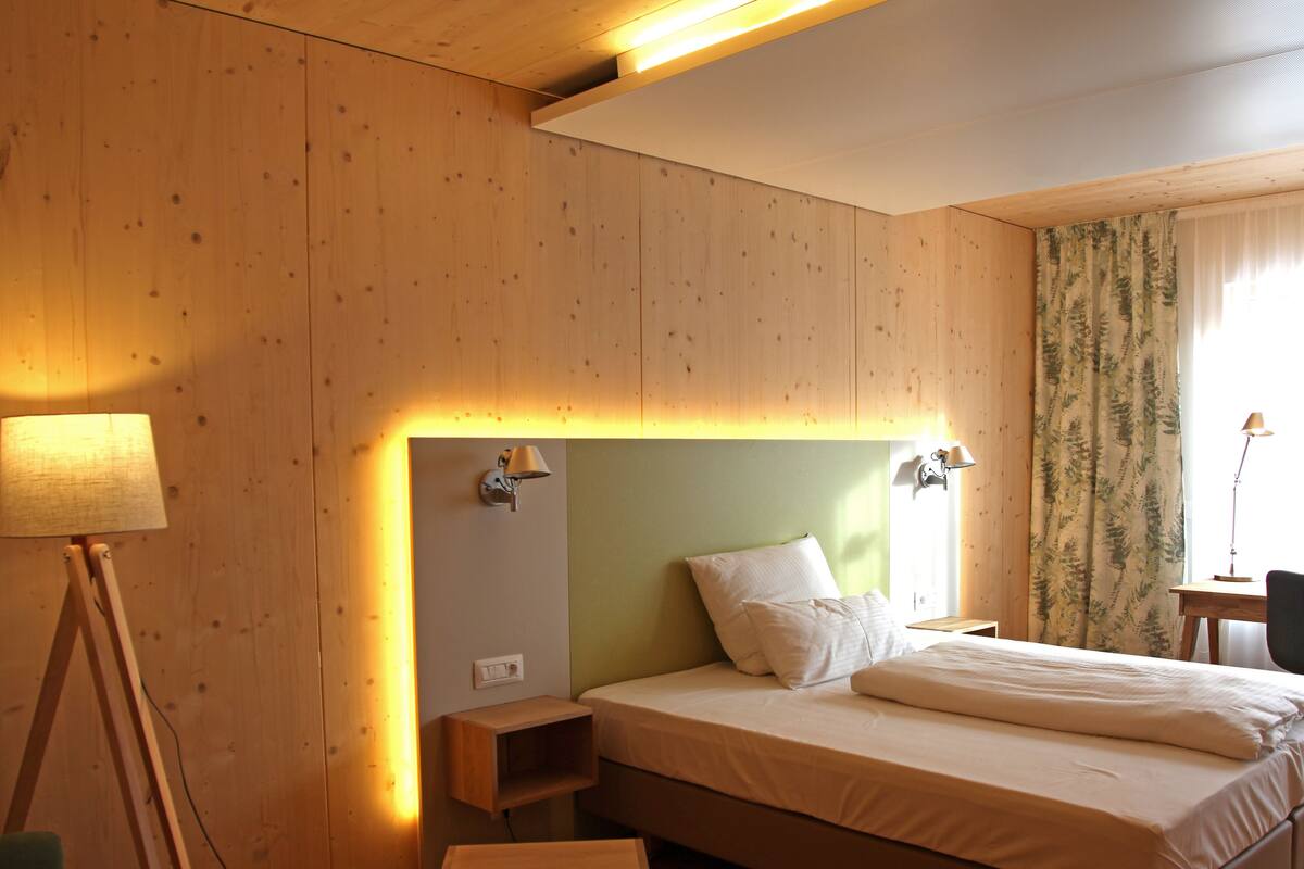 Ein Hotelzimmer mit Holzwand, einem Bett mit weißer Bettwäsche, einem Nachttisch, einer Lampe und einem Fenster mit Vorhängen.