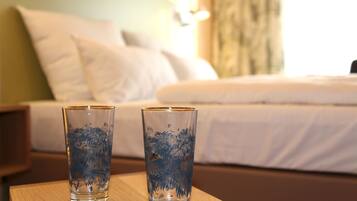 Dos vasos transparentes sobre una mesa de madera en una habitación de hotel, con una cama y una mesita de noche al fondo.