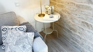 Um quarto acolhedor com sofá cinzento, almofadas estampadas, uma mesa redonda pequena com abajur, objetos decorativos e uma parede de tijolos.