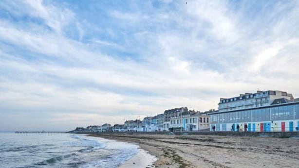 Um hotel à beira-mar com uma extensa faixa de areia, um cais que se projeta para o mar e uma fileira de edifícios coloridos ao longo da costa.
