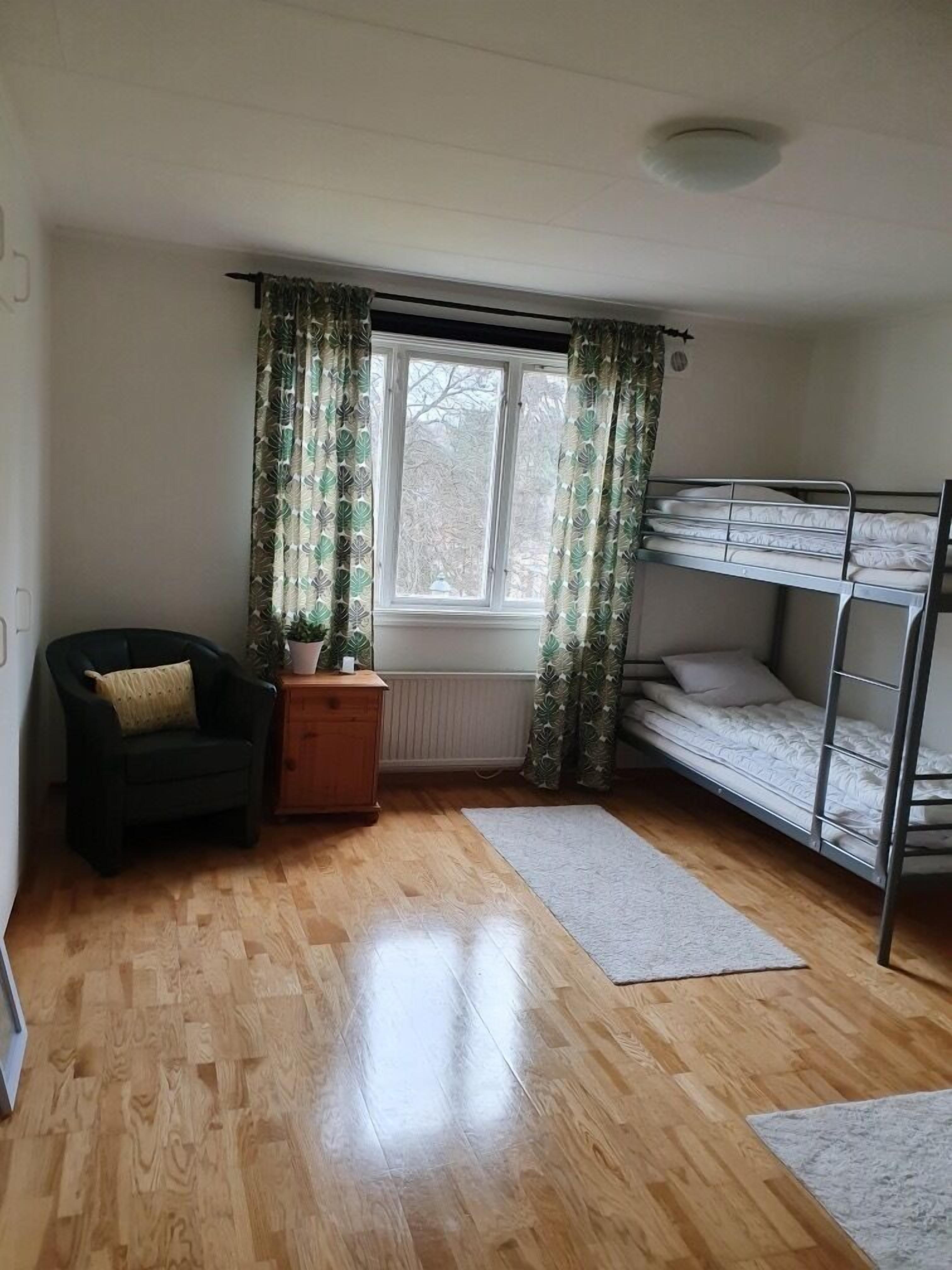 A room with a bunk bed, a chair, a small table with a plant, and a window with curtains.