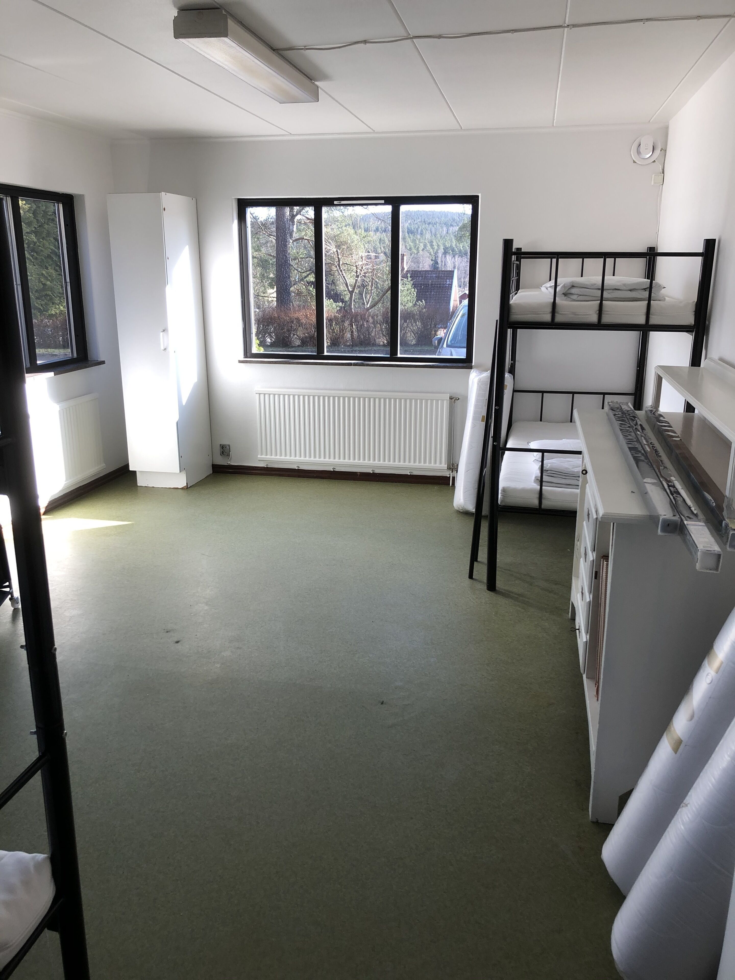 A room with bunk beds, a radiator, a window with a view of trees, and a white cabinet.