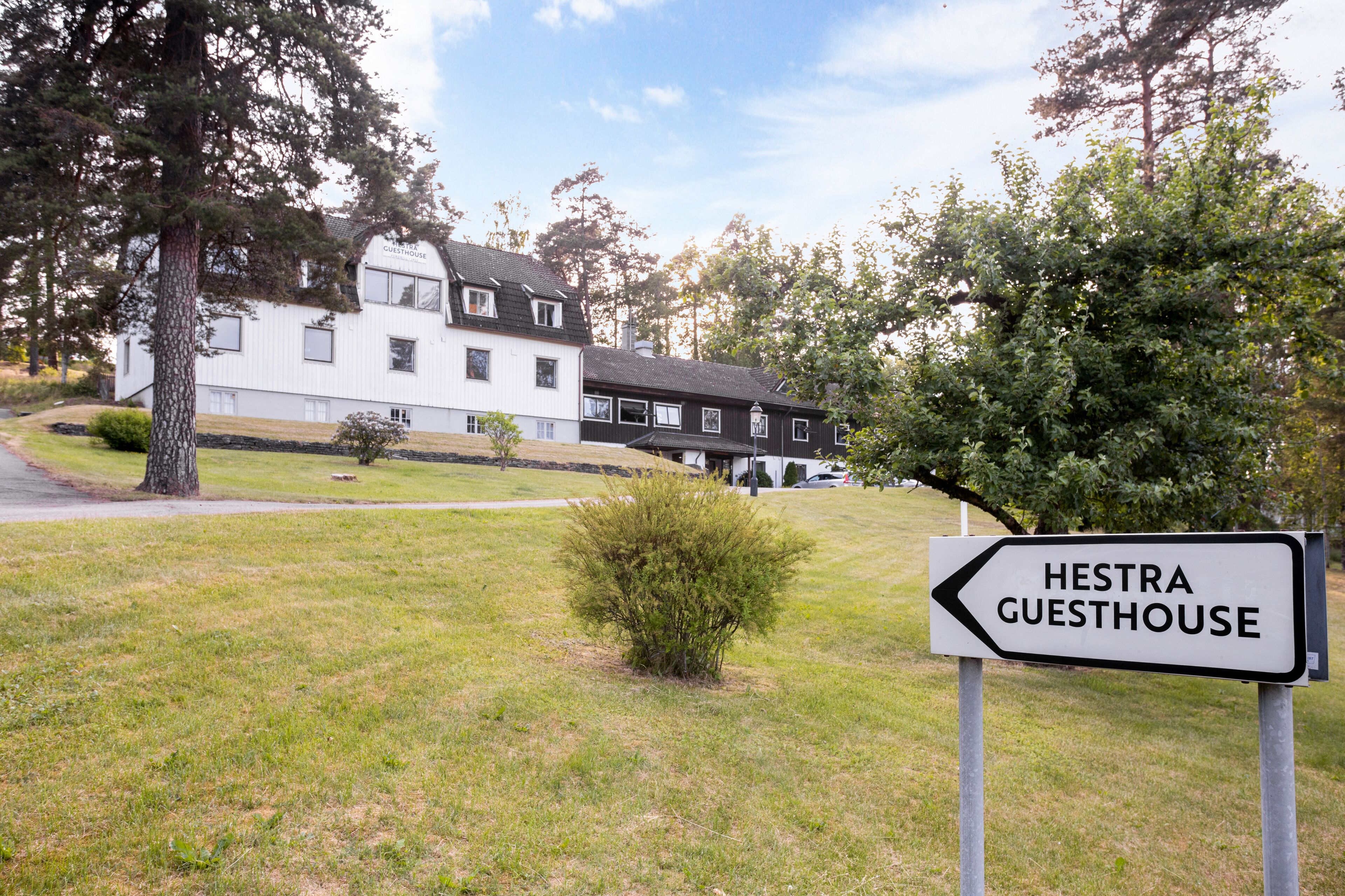 A sign for Hestra Guesthouse with a white building in the background.