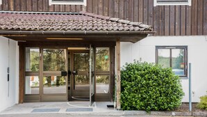 A building entrance with a tiled roof, glass doors, and a small green bush beside it.
