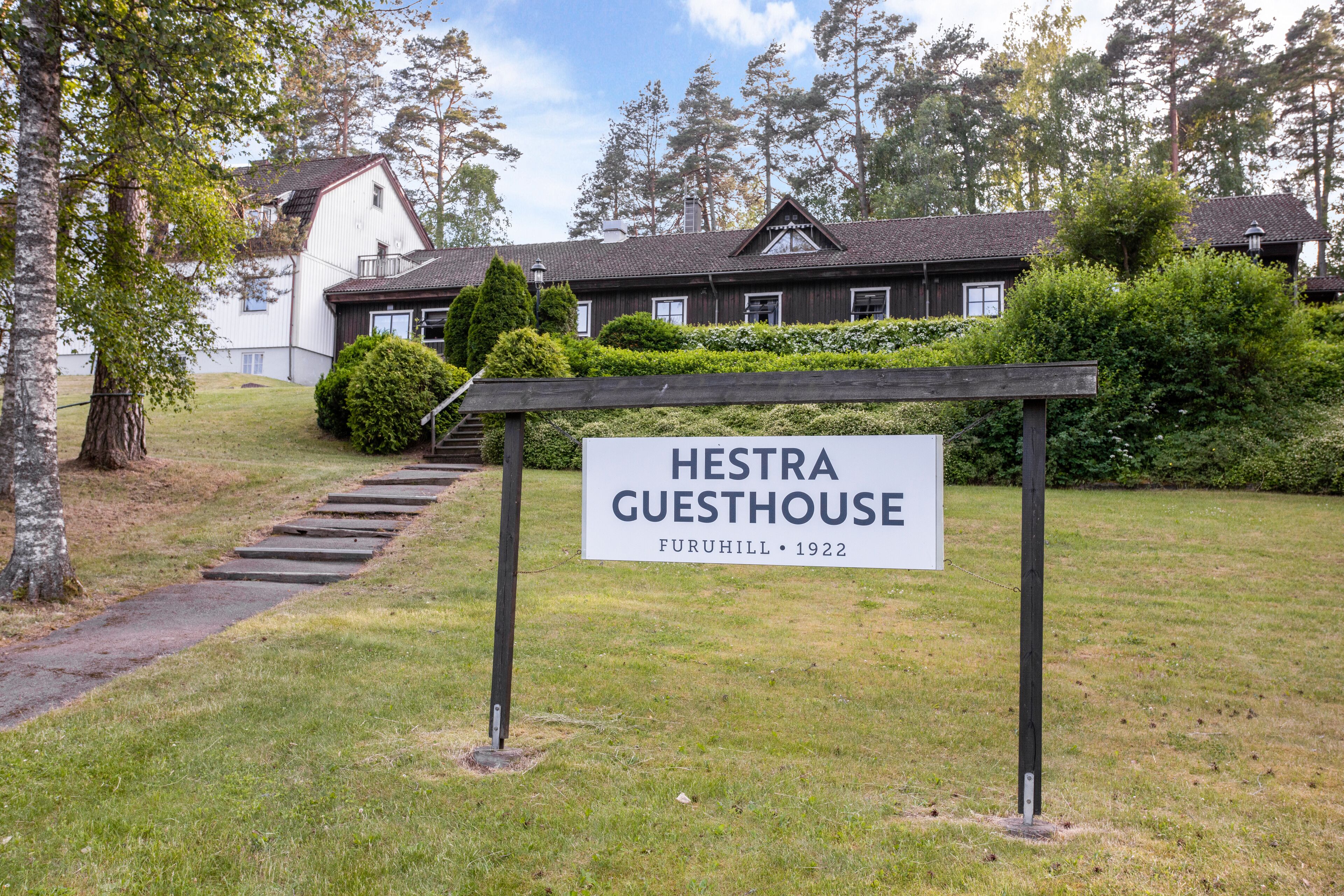A sign for Hestra Guesthouse in Furuhill, established in 1922, with a staircase leading to a building surrounded by greenery.